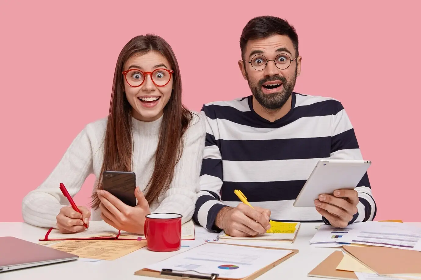 Des membres d’équipe sont assis à un même bureau, prennent des notes dans un carnet, se préparent à un séminaire, tiennent un smartphone moderne et une tablette tactile, affichent un regard heureux, examinent des documents avec des graphiques et sont engagés dans le processus de travail.